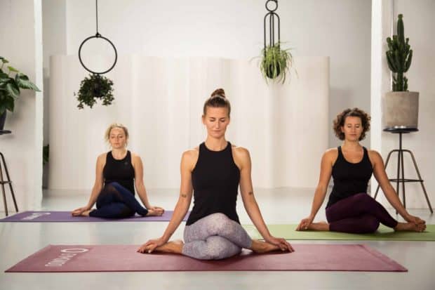 3 junge Frauen sitzen in hellem Raum auf bunten Yogamatten im Schneidersitz und geschlossenen Augen machen Yin Yoga