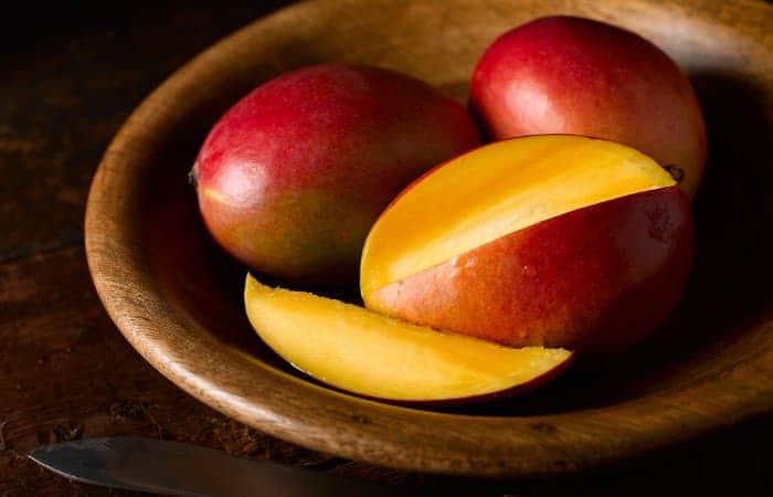 Eine in Scheiben geschnittene Mango, dahinter zwei ganze, rote Mangos