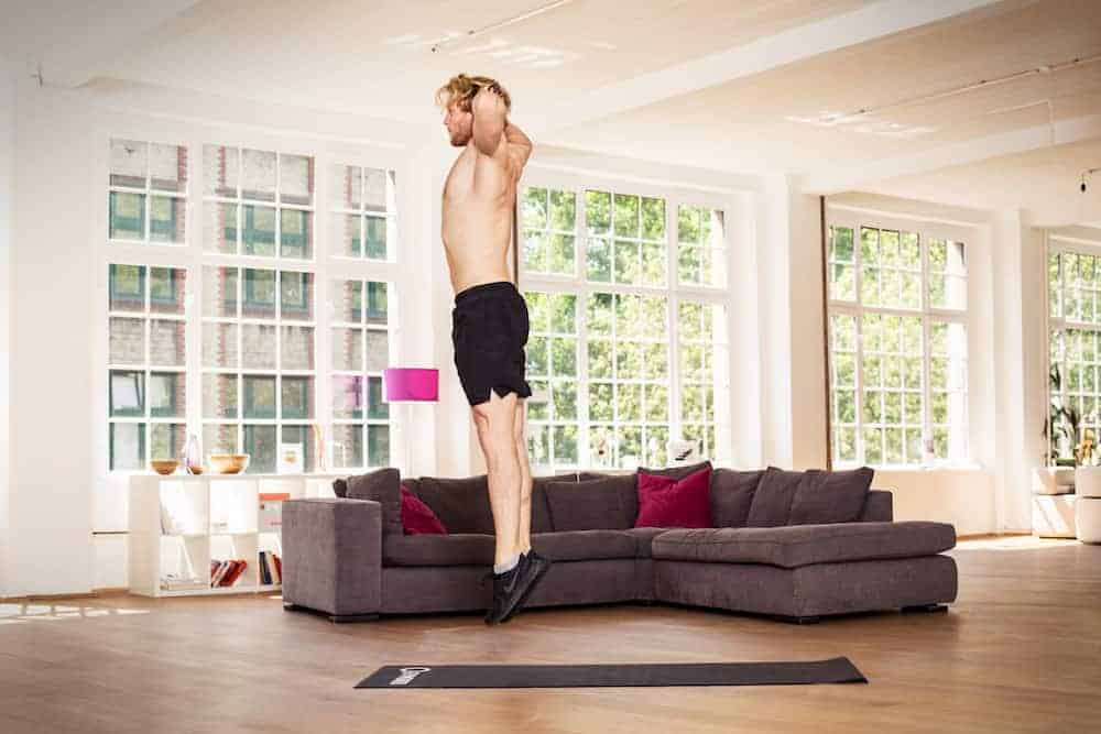 Mann in heller Wohnung auf dunkler Trainingsmatte macht einen Burpee und einen Strecksprung mit Händen hinter dem Kopf