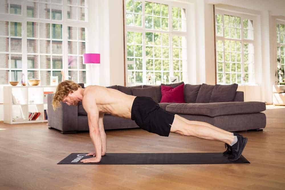 Mann in Liegestützposition auf dunkler Trainingsmatte in heller Wohnung macht Push Up