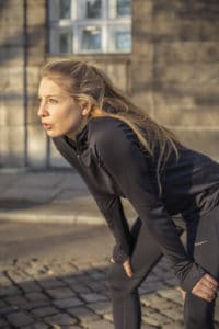 Frau beim Lauftraining in Laufkleidung atmet durch, die Hände auf die Knie gestützt.