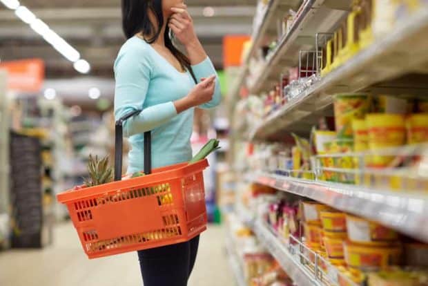 Frau mit Einkaufskorb zwischen den Regalen eines Supermarktes überlegt welche Lebensmittel sie kaufen soll