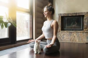 Junge Frau sitzt auf Yogamatte im Lotussitz daneben eine kleine Katze